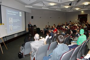 A large auditorium full of people, facing a screen and man at a lecturn