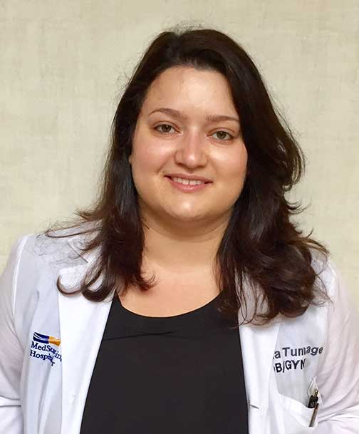 Irina Tunnage smiles, wearing a white coat with her name and “OB/GYN” embroidered on the right side