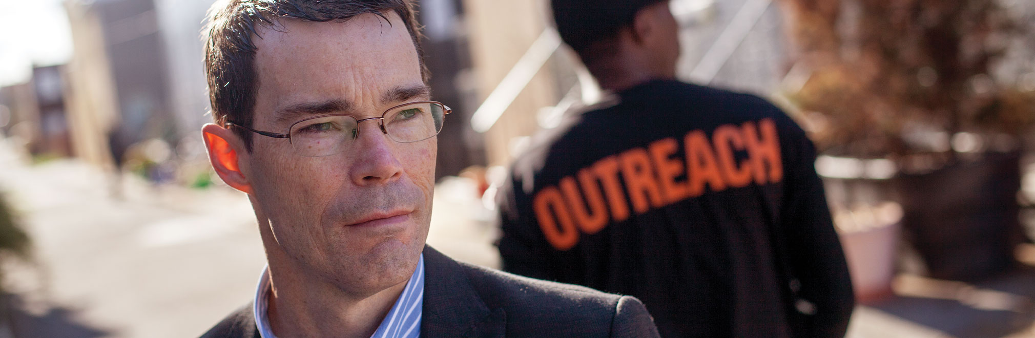 Person in suit behind person with a jacket that reads outreach.