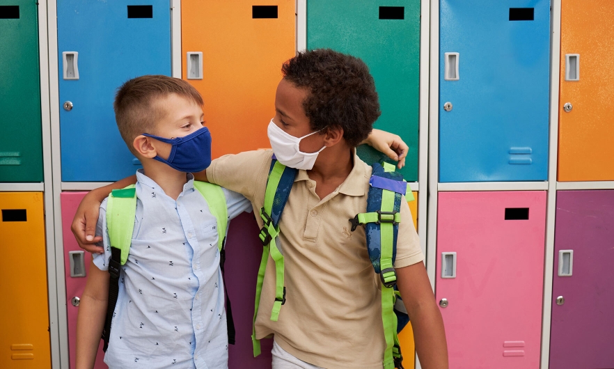 Two kids wearing masks embracing in front of school lockers