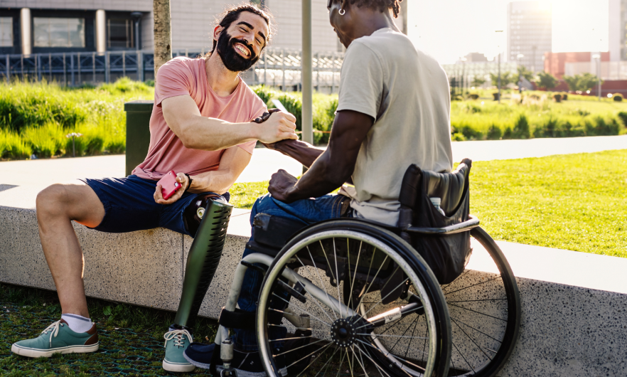 Man in wheelchair shakes hands with a smiling man with a prosthetic leg seated on stone bench outside