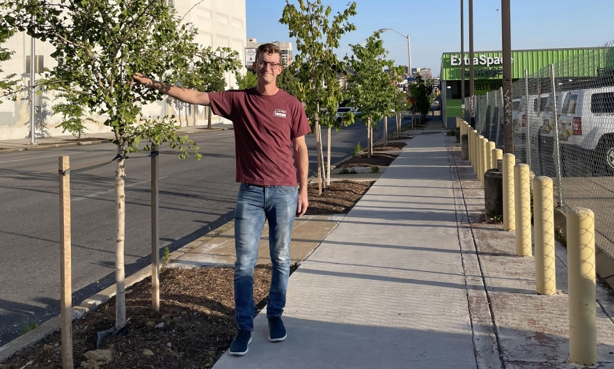 Man in red shirt and jeans standing on a sidewalk with his hand on a tree