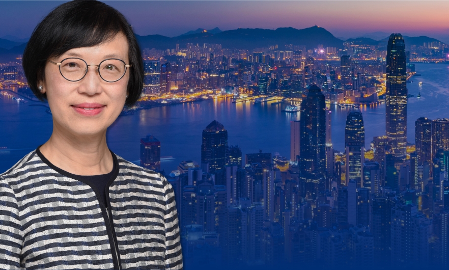 Photo of Dr. Chan, a smiling woman with short parted brown hair, brown eyes, and a light skin complexion, wearing eyeglasses and a black and white striped shirt, posing in front of a photo of the Hong Kong city skyline at night