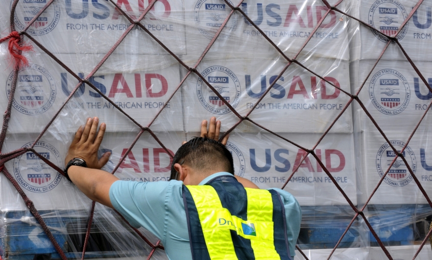 Worker pushes hands against a pallet stacked with boxes of USAID supplies
