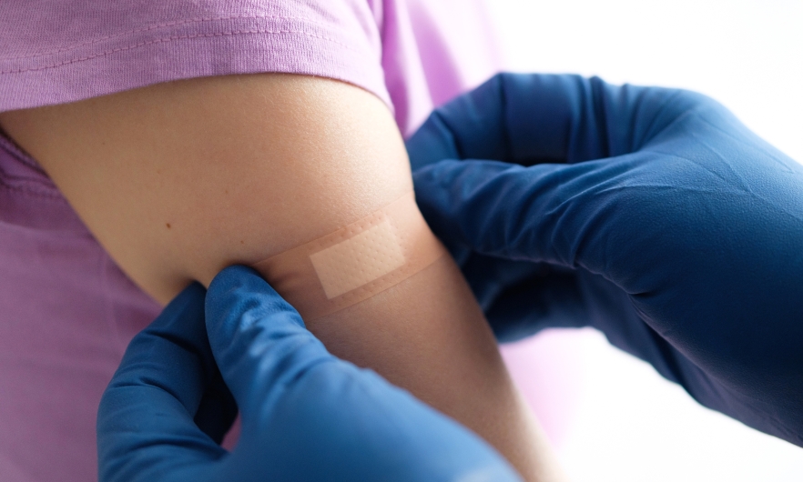 Hands wearing surgical gloves place bandaid on young child's upper arm.