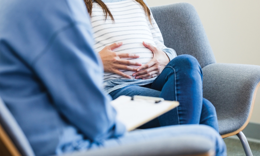 An unrecognizable expectant mom sitting in a chair puts her hands on her belly as she shares her worries about her baby with the unrecognizable therapist. 