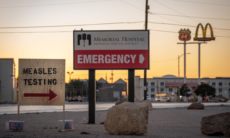 A plywood sign reads "Measles Testing" and points right. Behind it, a larger permanent sign points right towards the Memorial Hospital Emergency Department.