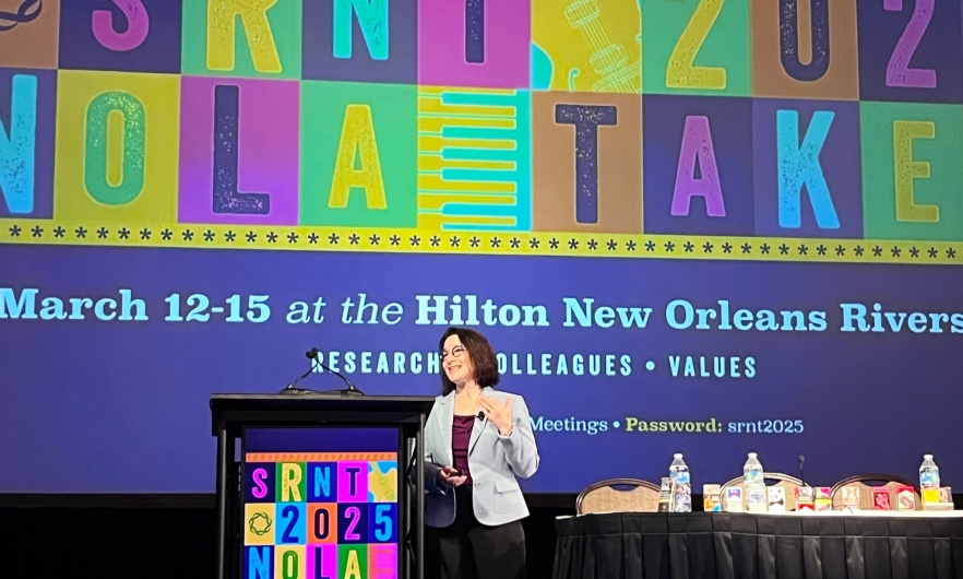 A woman in a blazer stands on stage in front of a colorful backdrop giving a presentations