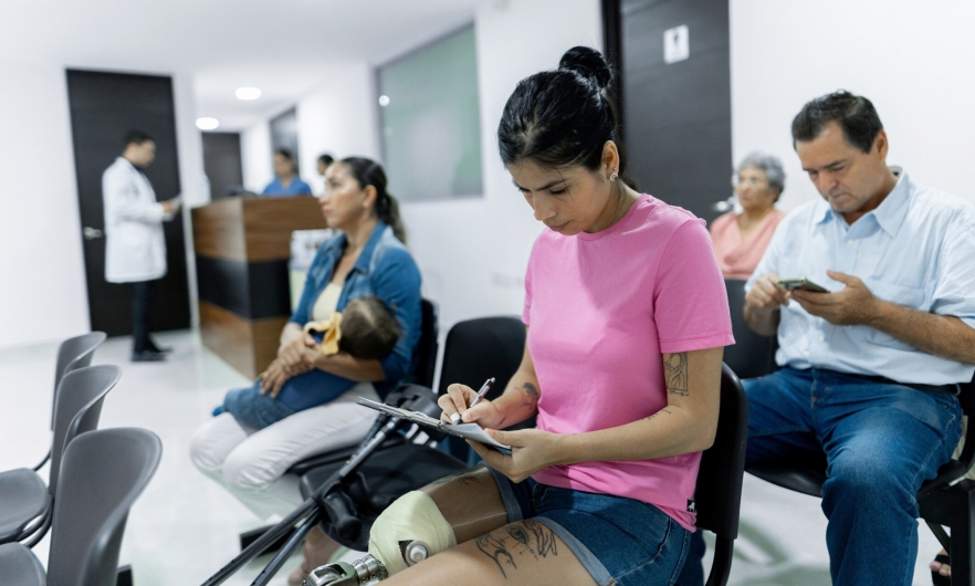 A woman with a prosthetic leg fills out forms in the waiting room of a medical office. A mother and baby, an older woman, and a middle-aged man also sit waiting.