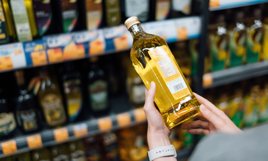 In front of shelves lined with various types of cooking oil, a shopper reads the label of a bottle of cooking oil.