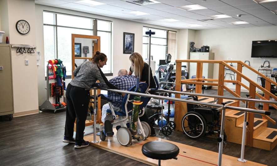 In a physical therapy office, two female therapists, one on either side, work with a man in a wheelchair who is navigating between parallel bars.