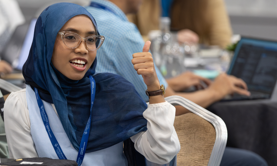 A woman wearing a blue hijab and glasses gives a thumbs up