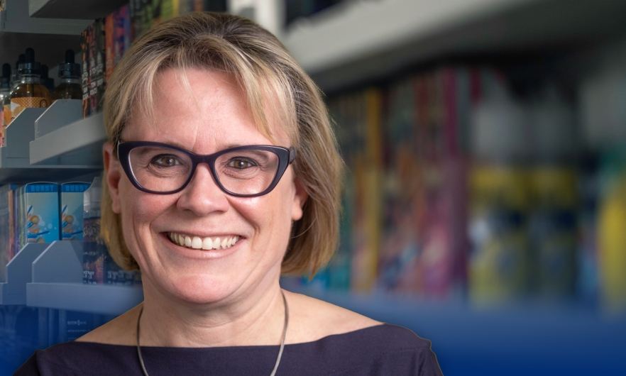 Katherine Clegg Smith, a woman with a light complexion, brown eyes, a friendly smile, dark framed eyeglasses, and neck-length blondish-brown hair, wearing a dark purple dress and silver necklace, standing in front of a blurry display of e-cigarette products on a store shelf