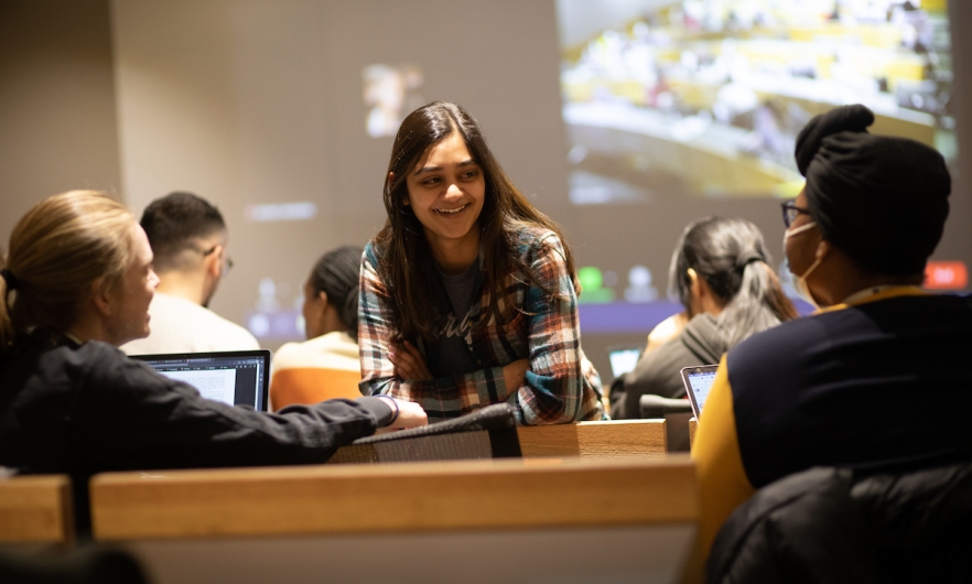 three students chat before class in a lecture hall set up for hybrid learning