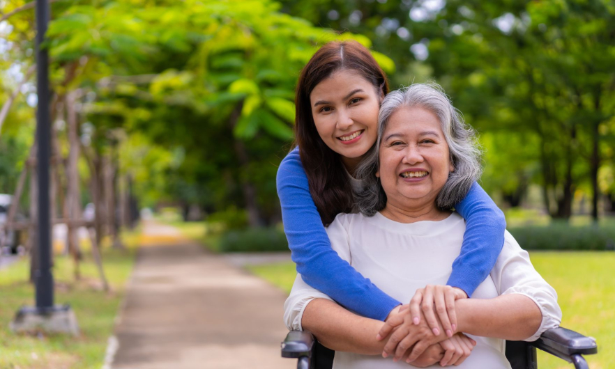 An elderly woman seated in a wheelchair is hugged from behind by a younger woman. The two smile at the camera.
