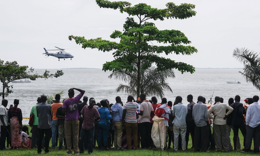 Dozens of bystanders look on as they watch rescuers in a helicopter search the site of a capsized cruise boat on Lake Victoria near Mutima village, south of Kampala, Uganda, on a grey, cloudy day.