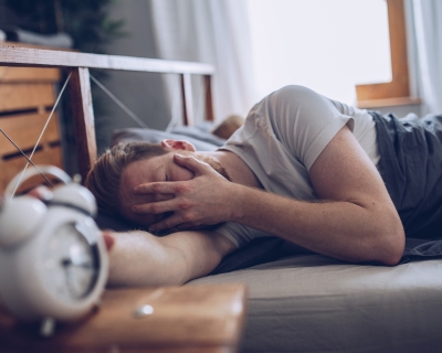 Caucasian male laying in bed covering face reaching for alarm clock