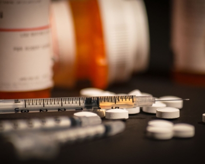 White pills are scattered on the counter near a few orange pill bottoms. A syringe is next to the scattered pills.