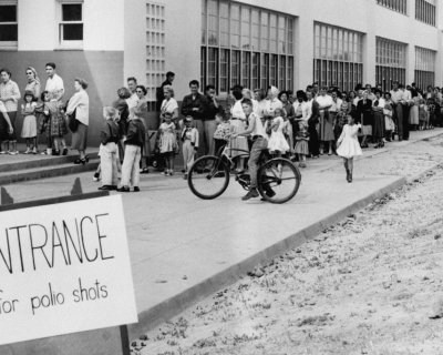 Black and white historical photo from 1955 of families lined up at a school for polio shots. The line wraps around the building.