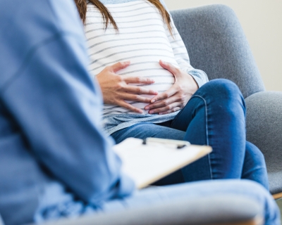 An unrecognizable expectant mom sitting in a chair puts her hands on her belly as she shares her worries about her baby with the unrecognizable therapist. 