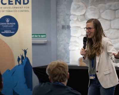 Graziele Grilo, a woman with a medium complexion and long brown hair, wearing eyeglasses and a white jacket, speaks to a room of young participants in a tobacco control leadership program