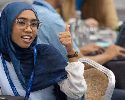 A woman wearing a blue hijab and glasses gives a thumbs up