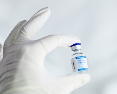 A white-gloved hand holds a vaccine vial with a blue and white label that reads "mRNA vaccine"