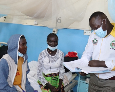 A doctor, standing and looking over a chart, attends to a patient seated on a cot.
