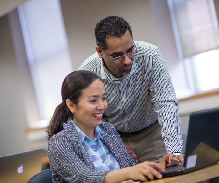 Man standing and woman sitting, both looking at laptop screen.