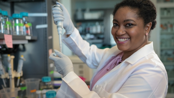 A woman in a lab with lab equipment in her hands smiling at the camera