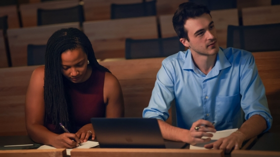 Two students sitting next to each other in classroom. One is looking to the side and the other is writing in a notebook.