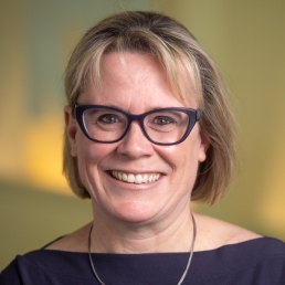 Headshot of Katherine Clegg Smith, a woman with a light complexion, brown eyes, a friendly smile, dark framed eyeglasses, and neck-length blondish-brown hair, wearing a dark purple dress and silver necklace, posed in front of a blurred background with warm tones