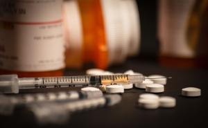 White pills are scattered on the counter near a few orange pill bottoms. A syringe is next to the scattered pills.