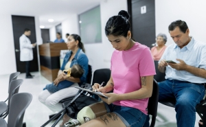 A woman with a prosthetic leg fills out forms in the waiting room of a medical office. A mother and baby, an older woman, and a middle-aged man also sit waiting.
