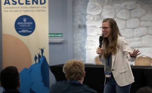 Graziele Grilo, a woman with a medium complexion and long brown hair, wearing eyeglasses and a white jacket, speaks to a room of young participants in a tobacco control leadership program