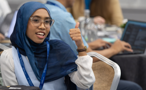A woman wearing a blue hijab and glasses gives a thumbs up