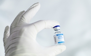 A white-gloved hand holds a vaccine vial with a blue and white label that reads "mRNA vaccine"