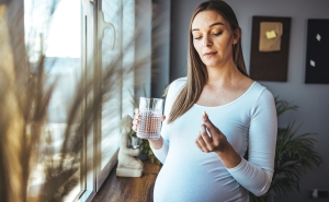 A pregnant woman with long brown hair wearing a white shirt holds a pill in one hand and a glass of water in the other. She is standing by some windows in her living room.