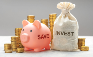 A pink piggy bank and a white sack sit side by side in front of a stack of coins. The piggy bank says save while the sack says invest.