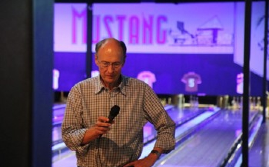 Roger McMacken stands holding a microphone, with the Mustang bowling lanes behind