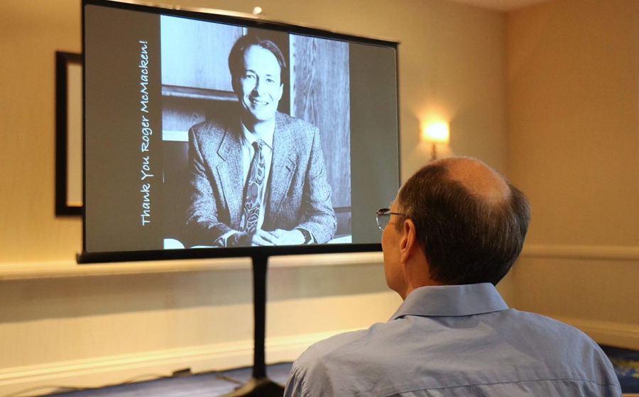 Roger McMacken sits in the front row, facing a screen with much younger photo of him in black and white and the message Thank you Roger McMacken!