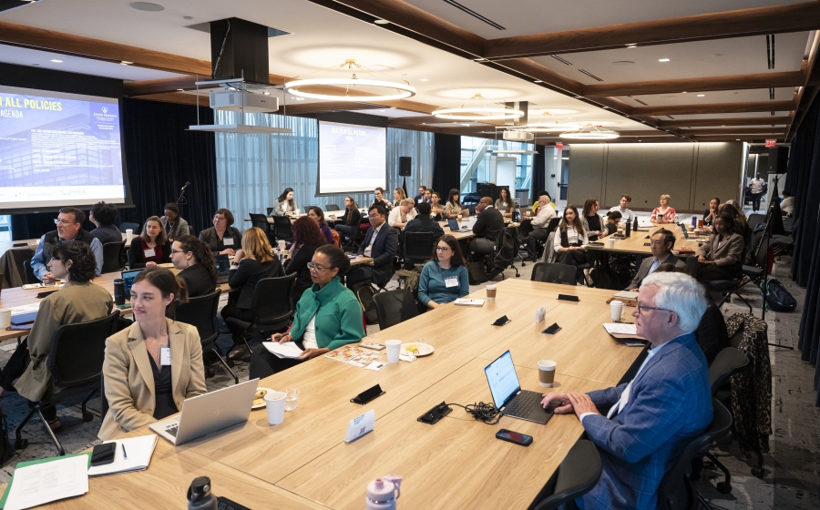 A racial and gender diverse group of people seated at several long wooden tables, some with laptops or coffee cups by their arms. Most are facing towards the front of the wide room.