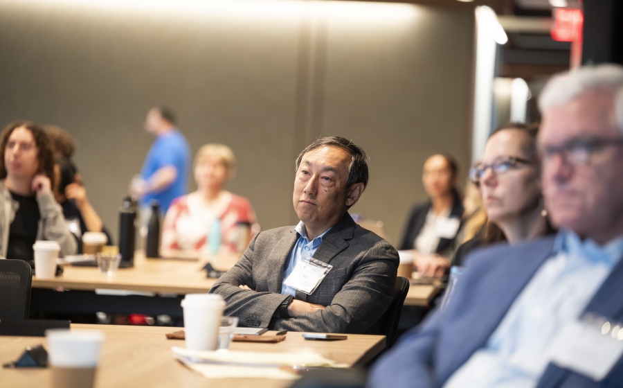 A man in a black suit jacket is seated at a wooden table with a coffee cup near him. His attention is towards front of the room. In the background and foreground, multiple people at other tables are facing the same direction.