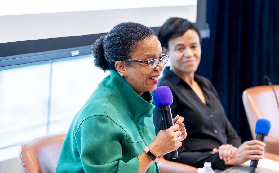 In the focused foreground, Maryland State Delegate Robbyn T. Lewis smiles as she speaks into a hand-held microphone. In the unfocused background, Department Chair Keshia Pollack Porter smiles in Lewis' direction.