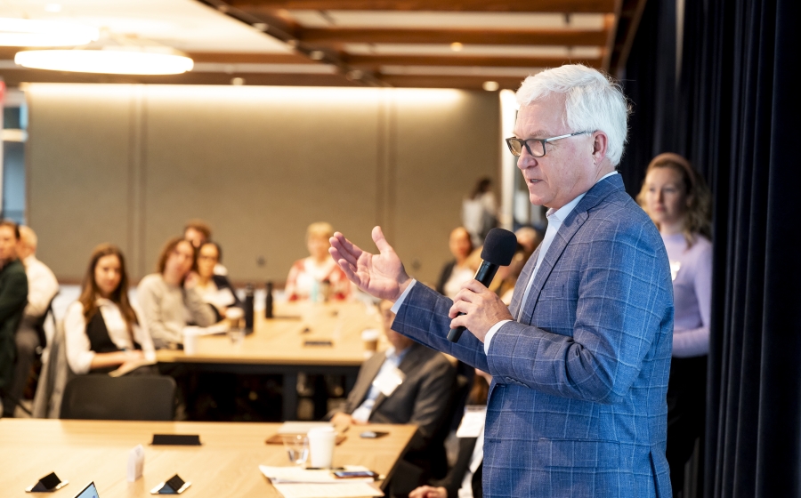 A man wearing a dark grey suit jacket stands next to a wooden table while holding a hand-held microphone. He's speaking towards the front of the room. In the unfocused background, multiple people are seated at a wooden table.