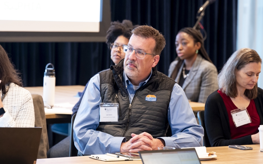 A man in a light blue dress shirt with a black vest over it is seated at a wooden table. His attention is towards front of the room, which is out of frame.