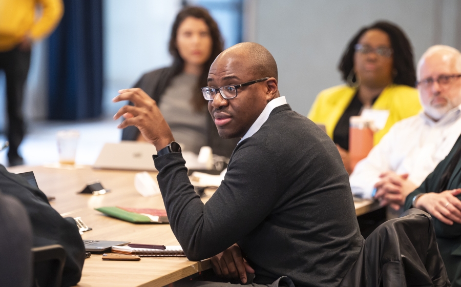 A man in a black sweater is seated at a wooden table. His attention is towards front of the room as he gestures with his hand. The front of the room is out of frame.