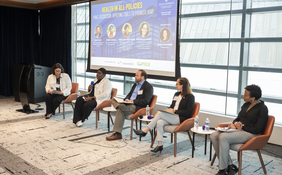 Four women and one man are seated in a row at the front of the room with a projector screen behind them. One of the women, Danay Hough, is holding a microphone as she speaks. The screen is entitled Health in All Policies Panel Discussion: Applying Tools to Promote HiAP. The panelists are listed as Jimmy Dills, Danaya Hough, Keshia Pollack Porter, and Elizabeth A. Ramey. The moderator is listed as Kristin Robinson.