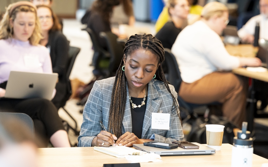 A woman in a black shirt and grey suit jacket is seated at a wooden table. She's writing onto a white piece of paper. Behind her at other tables, people are typing into their laptops or writing onto paper.
