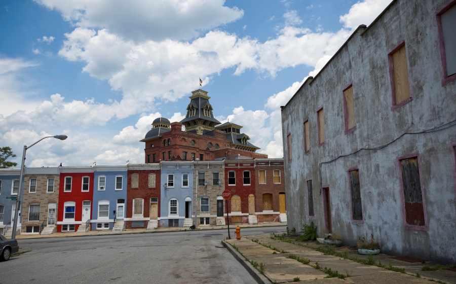 Street view of baltimore rowhouses, many with boarded up windows; rising behind them is them a Victorian-era brick building with elaborate decrative roofs